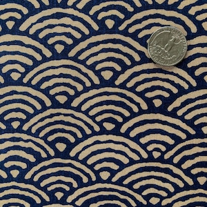 Può includere: Primo piano di un tessuto con un motivo a onde ripetute in blu navy e beige. Una moneta è posta sul tessuto per la scala. Il design ricorda l'arte tradizionale giapponese.