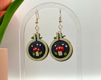 May include: A pair of handmade earrings featuring miniature embroidered mushroom designs. Each earring is set in a small wooden embroidery hoop with a dark blue background, a red mushroom, and green leaves. The earrings hang from gold-colored ear wires.