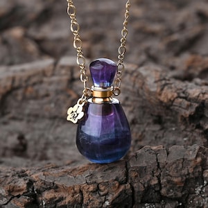 Puede incluir: Un collar de cadena dorada con un peque&ntilde;o colgante en forma de l&aacute;grima. El colgante es un frasco de vidrio con un esquema de color p&uacute;rpura y azul. Un peque&ntilde;o dije dorado con la letra "M" cuelga del frasco.
