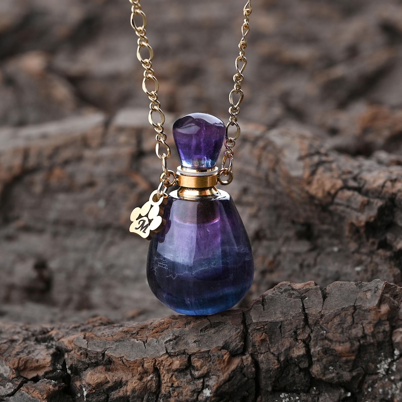 Puede incluir: Un collar de cadena dorada con un peque&ntilde;o colgante en forma de l&aacute;grima. El colgante es un frasco de vidrio con un esquema de color p&uacute;rpura y azul. Un peque&ntilde;o dije dorado con la letra "M" cuelga del frasco.