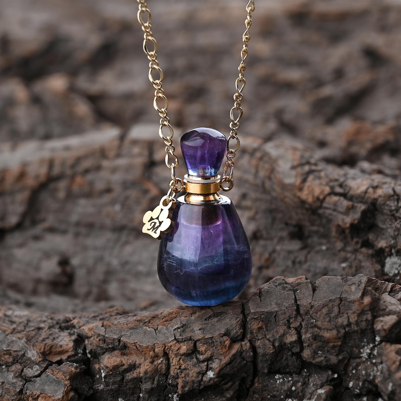 Puede incluir: Un collar de cadena dorada con un peque&ntilde;o colgante de botella de perfume. La botella es una mezcla de tonos morados y azules, con una tapa dorada y un peque&ntilde;o dije. El fondo es una superficie marr&oacute;n texturizada.