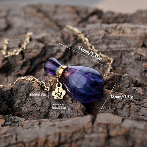Puede incluir: Un collar morado y dorado con un colgante en forma de peque&ntilde;o frasco de perfume. El colgante mide 34 mm de largo y 18 mm de ancho, con una cadena dorada y un charm de huella de pata. El charm tiene la letra "M".