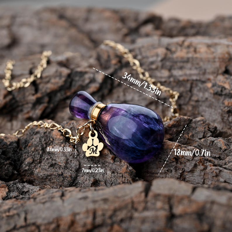 Puede incluir: Un collar morado y dorado con un colgante en forma de peque&ntilde;o frasco de perfume. El colgante mide 34 mm de largo y 18 mm de ancho, con una cadena dorada y un charm de huella de pata. El charm tiene la letra "M".