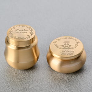 Könnte beinhalten: Zwei goldfarbene Urnen. Eine ist höher mit einem floralen Design und dem Namen "Luctken" und den Jahren "2015-2025" auf dem Deckel eingraviert. Die andere Urne ist kürzer, mit den Worten "In Loving Memory" und einem Pfotenabdruck-Design, ebenfalls mit dem Namen "Luciken" und den Jahren "2015-2025".