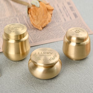 May include: Three small, gold-colored urns with engraved inscriptions. One urn reads "Lucieen 2015-2024" within a laurel wreath. Another says "LUCKY 2015-2024" with a heart and leaves. The third urn has a paw print and the words "In Loving Memory".