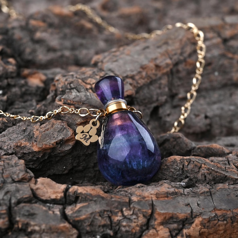 Puede incluir: Un collar de cadena dorada con un colgante en forma de l&aacute;grima. El colgante es una piedra de color p&uacute;rpura y azul oscuro, con una tapa dorada y un peque&ntilde;o dije con una huella de pata y la letra "M". El collar se muestra sobre una superficie marr&oacute;n texturizada.