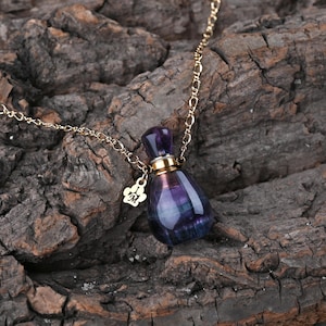 Puede incluir: Un collar de cadena dorada con un peque&ntilde;o colgante de botella de perfume de cristal morado y azul. La botella tiene una tapa dorada y un peque&ntilde;o dije con una huella de pata y la letra "M".