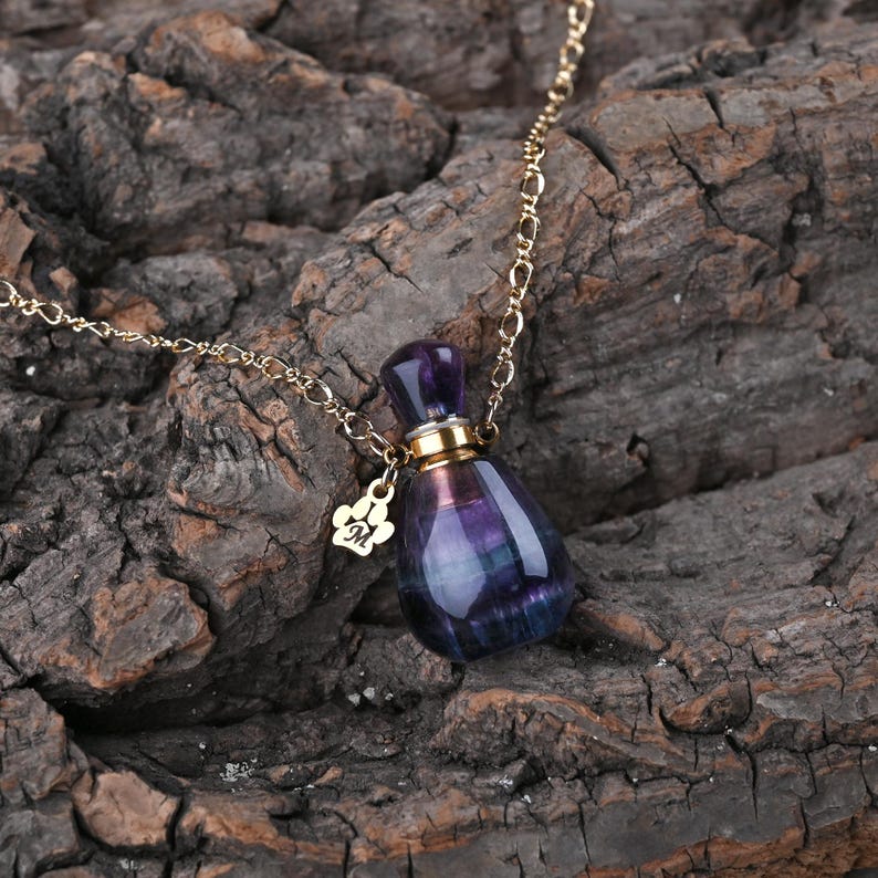 Puede incluir: Un collar de cadena dorada con un peque&ntilde;o colgante de botella de perfume de cristal morado y azul. La botella tiene una tapa dorada y un peque&ntilde;o dije con una huella de pata y la letra "M".