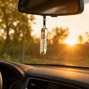 May include: A gold-toned metal car rearview mirror hanging ornament featuring a cylindrical glass vial with a wooden cap. The metal frame is engraved with the text Koda 2015-2026. The ornament is suspended by a black cord with a small gold bead.