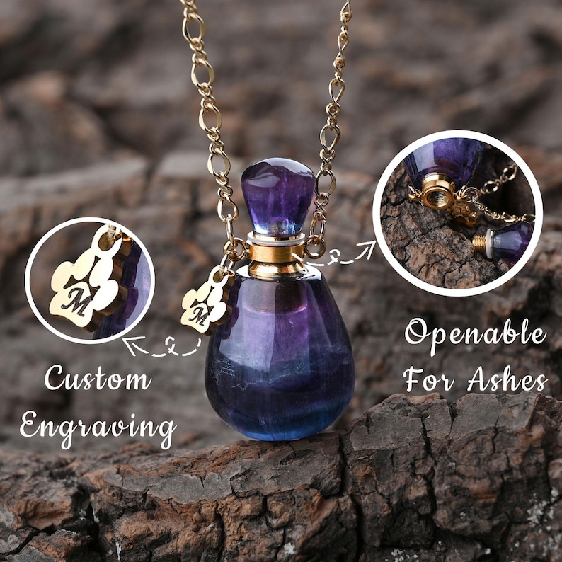 Puede incluir: Un collar con colgante de cristal morado y azul con una cadena dorada. El colgante es una peque&ntilde;a botella con una tapa dorada y un tap&oacute;n morado. Un dije dorado con forma de huella de pata con la letra "M" est&aacute; adjunto. El texto dice "Grabado personalizado" y "Apertura para cenizas."