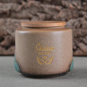 May include: A ceramic urn with a wooden lid. The urn is tan with teal accents and features the word "Shalom" in gold script, along with the years 2019-2025 and a gold angel wing design. The urn has a rounded shape.