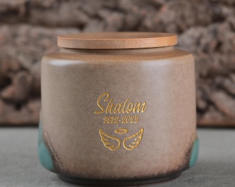 May include: A ceramic urn with a wooden lid. The urn is tan with teal accents and features the word "Shalom" in gold script, along with the years 2019-2025 and a gold angel wing design. The urn has a rounded shape.