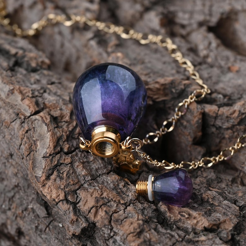 Puede incluir: Un colgante de piedra preciosa morada y azul en forma de l&aacute;grima, con una tapa dorada atornillable y un tap&oacute;n a juego. El colgante cuelga de una delicada cadena dorada, sobre una superficie de madera marr&oacute;n texturizada.