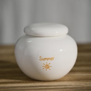 May include: A white ceramic jar with a lid, featuring the word "Summer" and a sun graphic in gold. The jar has a rounded shape and sits on a wooden surface.