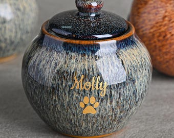 Może przedstawiać: Ceramiczna urna dla zwierząt z pokrywką. Urna jest nakrapiana na niebiesko i beżowo ze złotym odciskiem łapy i imieniem "Molly". Pokrywka ma ciemne, nakrapiane wykończenie z małym guzikiem. Urna jest okrągła.