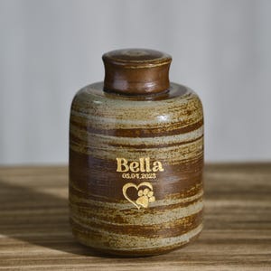 May include: A ceramic pet urn with a brown and beige striped pattern. The urn has a lid and features the name "Bella", the date "05.04.2023", and a paw print inside a heart.