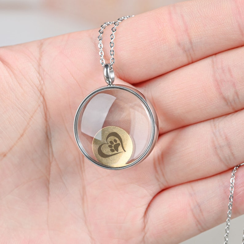 Pu&ograve; includere: Collana con catena d'argento con un ciondolo rotondo in vetro trasparente che contiene un cuore dorato con un'impronta di zampa all'interno.