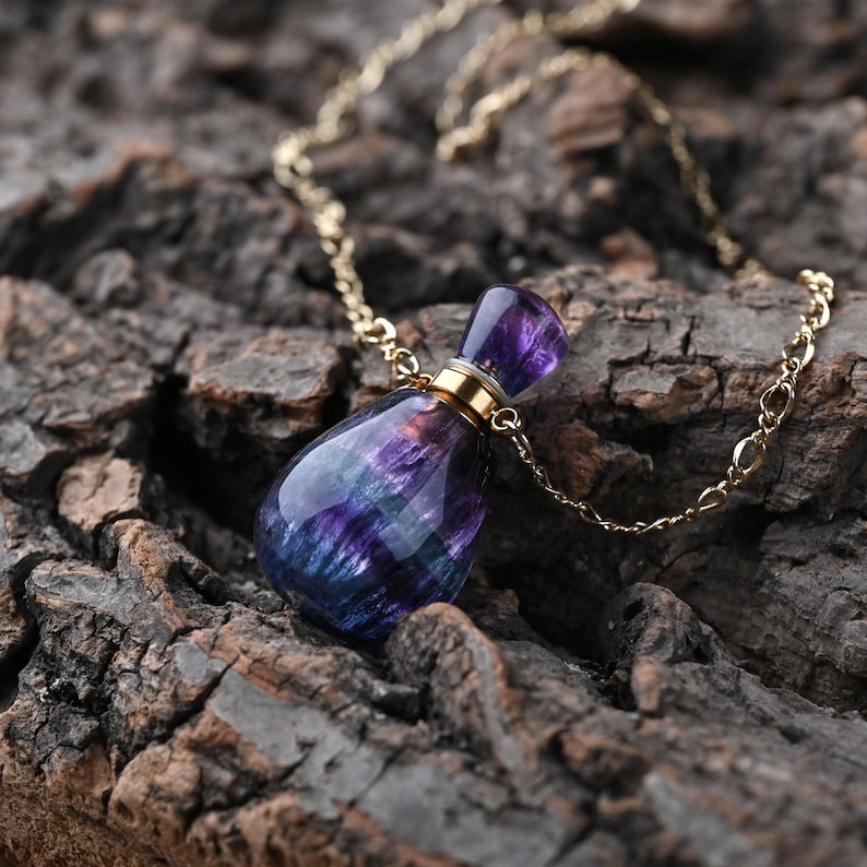Puede incluir: Collar con un colgante de un peque&ntilde;o frasco de perfume. El frasco, con forma de l&aacute;grima, est&aacute; hecho de una piedra transl&uacute;cida con tonos morados y azules. Tiene una tapa dorada y cuelga de una fina cadena dorada.