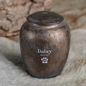 Könnte beinhalten: Eine kleine, dunkelbraune Keramikurne mit Deckel. Die Urne hat eine abgenutzte Oberfläche und ist mit dem Namen "Daisy" und dem Datum "2023/12" graviert. Ein Pfotenabdruck mit einem Herzen ist ebenfalls in die Urne graviert.