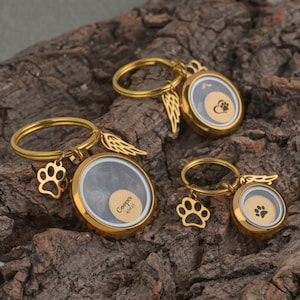 May include: Gold-colored pet memorial keychains. Each keychain features a round locket with a paw print or a small wing charm. One keychain has the name "Cooper" and the date "10/1/25" engraved on it.