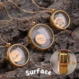 May include: Gold-colored locket necklaces with clear glass faces. One locket features a paw print, while others have personalized text. The necklaces are displayed on a textured, dark brown surface. The locket's circular design is visible.