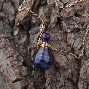 Puede incluir: Collar con colgante de una peque&ntilde;a botella en forma de l&aacute;grima de piedra morada y azul. La botella tiene una tapa dorada y cuelga de una fina cadena dorada. El fondo es una superficie marr&oacute;n texturizada.