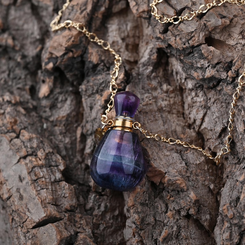 Puede incluir: Collar con colgante de una peque&ntilde;a botella en forma de l&aacute;grima de piedra morada y azul. La botella tiene una tapa dorada y cuelga de una fina cadena dorada. El fondo es una superficie marr&oacute;n texturizada.