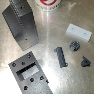 Könnte beinhalten: Eine Ansammlung dunkelgrauer 3D-gedruckter Komponenten, darunter ein rechteckiger Rahmen mit einem Ausschnitt, ein langes, abgewinkeltes Teil und mehrere kleinere Teile. Eine weiße Tasse mit dem Ghostbusters-Logo ist ebenfalls vorhanden.