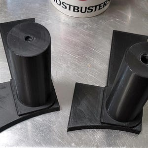 May include: Two black 3D-printed components, each with a cylindrical post and a rectangular base. The posts have a threaded hole at the top. The bases have a curved edge. The components are on a metallic surface.