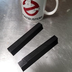 以下が含まれることがあります： 赤と白のゴーストバスターズのロゴが入った白いゴーストバスターズのマグカップ。テクスチャ加工された表面を持つ2つの黒い長方形のオブジェクトも画像にあります。