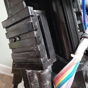 May include: Close-up of a black plastic component with a series of grooves and a slot. The component is attached to a larger black object with a bundle of colorful wires.