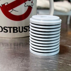 May include: A stack of light grey, circular, ridged objects. The objects are cylindrical and appear to be made of plastic. In the background is a white mug with the Ghostbusters logo and the word "GHOSTBUSTERS" in black.