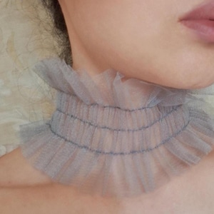 Könnte beinhalten: Ein grauer Tüll-Choker mit mehreren Lagen gerüschtem Stoff. Der Choker ist so konzipiert, dass er um den Hals getragen wird und ein zartes, luftiges Aussehen hat. Der Stoff hat eine weiche, gedämpfte graue Farbe.