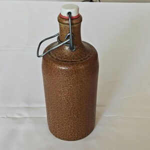 Könnte beinhalten: Eine braune Keramikflasche mit weißem Verschluss und Metallverschluss. Die Flasche hat eine strukturierte Oberfläche und eine zylindrische Form, die sich zum Aufbewahren von Flüssigkeiten eignet. Die Flasche ist etwa 25 cm hoch.