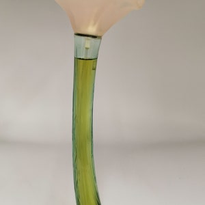 Könnte beinhalten: Eine Parfümflasche in Form einer Blume. Sie hat einen hellrosa Blütenkopf und einen langen, gebogenen grünen Stiel. Die Flüssigkeit im Inneren hat einen hellgelb-grünen Farbton. Die Flasche steht vor einem weißen Hintergrund.