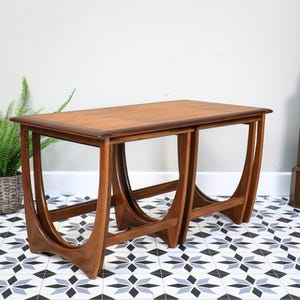 May include: A set of two nesting tables crafted from medium-toned wood. Each table features a rectangular top with rounded edges and distinctive U-shaped side supports that curve inward, creating a continuous frame design for the base.