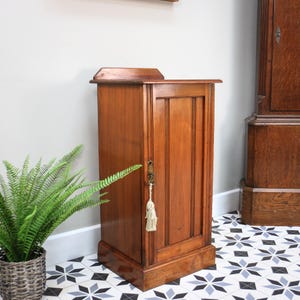 May include: A tall, rectangular wooden cabinet with a medium brown finish. The cabinet features a single vertical door with a recessed panel design, a brass keyhole escutcheon, and a decorative cream-coloured fabric tassel hanging from the key.