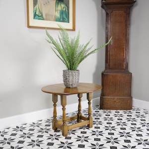 May include: A small, round wooden table with a natural finish. It features four turned legs and a hinged top. A woven grey pot with a green plant sits on the table. A tall wooden grandfather clock is in the background.