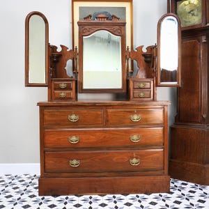 May include: A brown wooden vanity dresser featuring a large central mirror flanked by two smaller, adjustable side mirrors. The unit includes two small drawers on each side and three full-width drawers below, all accented with ornate brass hardware.