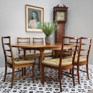 May include: A mid-century dining set featuring a round wooden table and four chairs with light brown leather cushions. A framed portrait and a tall grandfather clock are in the background. A potted plant sits on the table.