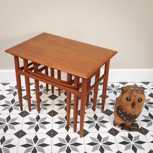 May include: A set of three wooden nesting tables with a rectangular top and slender, tapered legs. The tables are a warm brown colour and have a mid-century modern design. An owl figurine is next to the tables.