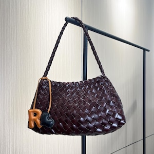 May include: A brown leather woven handbag with a black heart and orange letter R charm.