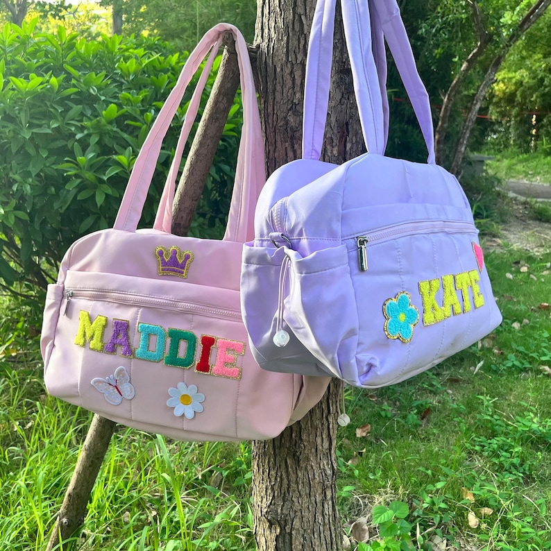 May include: Two personalized tote bags in pink and lavender hang from a tree. The pink bag has the name "MADDIE" in colorful letters, a butterfly, and a flower. The lavender bag has the name "KATE" and a flower.