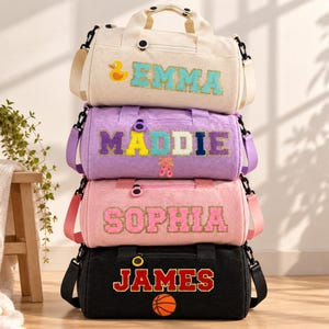 May include: A stack of four corduroy duffel bags in cream, purple, pink, and black. Each bag features a personalized name in varsity-style letters, including EMMA with a duck, MADDIE with ballet slippers, SOPHIA, and JAMES with a basketball.