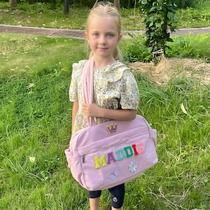 May include: A light pink shoulder bag with the name "MADDIE" in colorful embroidered letters. The bag features a gold crown, a butterfly, and a flower patch. The bag has a zipper closure and a shoulder strap.