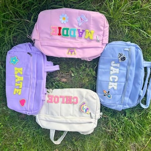 May include: Four quilted backpacks in pastel colors: pink, purple, white, and blue. Each backpack features a name patch in a contrasting color, with decorative patches like flowers, butterflies, and a unicorn. The backpacks are on a bed of green grass.