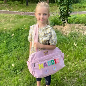 May include: A light pink shoulder bag with the name "MADDIE" in colorful embroidered letters. The bag features a gold crown, a butterfly, and a flower patch. The bag has a zipper closure and a shoulder strap.