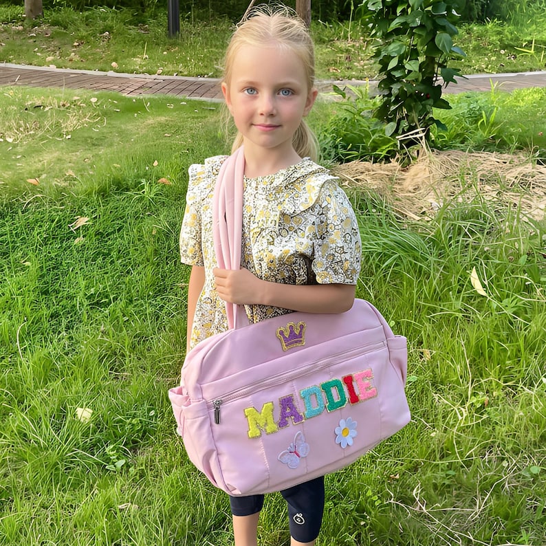 May include: A light pink shoulder bag with the name "MADDIE" in colorful embroidered letters. The bag features a gold crown, a butterfly, and a flower patch. The bag has a zipper closure and a shoulder strap.