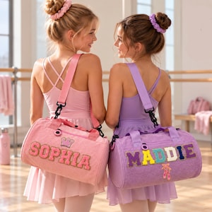 Op de afbeelding: Twee cilindervormige sporttassen van corduroy stof. De roze tas heeft de naam SOPHIA in glitterletters met een kroonicoon. De paarse tas heeft de naam MADDIE in kleurrijke letters met een icoon van balletschoenen.