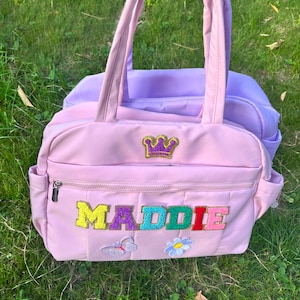 May include: Pink quilted tote bag with the name "MADDIE" in colorful letters. Features a gold crown patch and a butterfly and flower applique. A lavender bag is visible behind it.